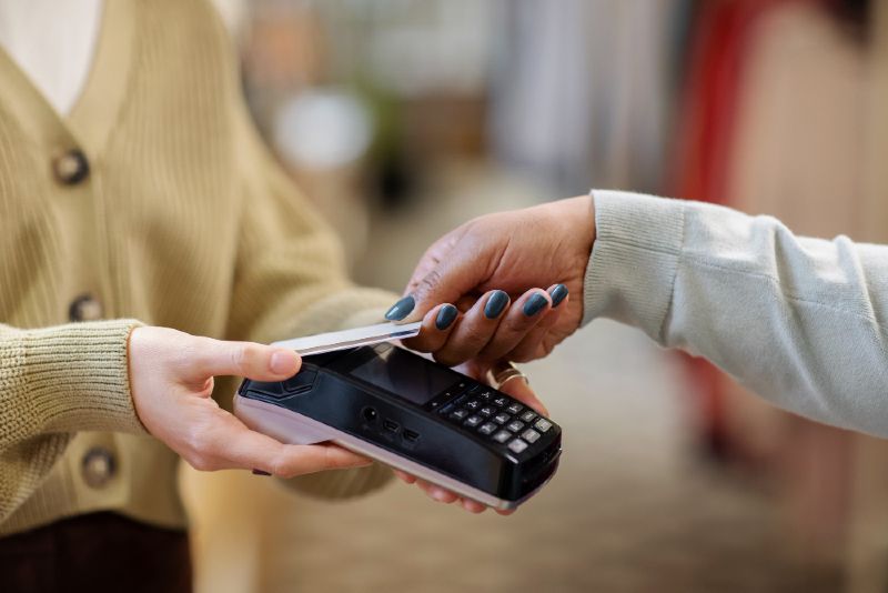 A mão de uma mulher segura um cartão de crédito próximo a uma máquina de cartão segurado por uma outra pessoa com as duas mãos