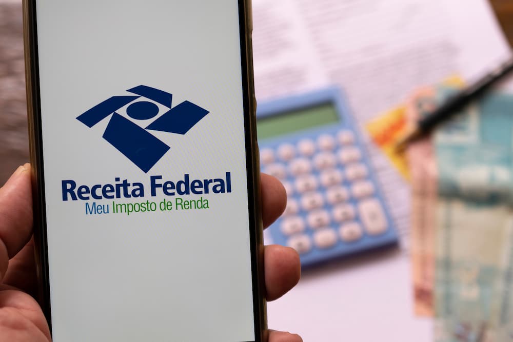 Uma pessoa segurando um celular com o logo da Receita Federal na tela. Ao fundo uma mesa com papéis e cédulas.