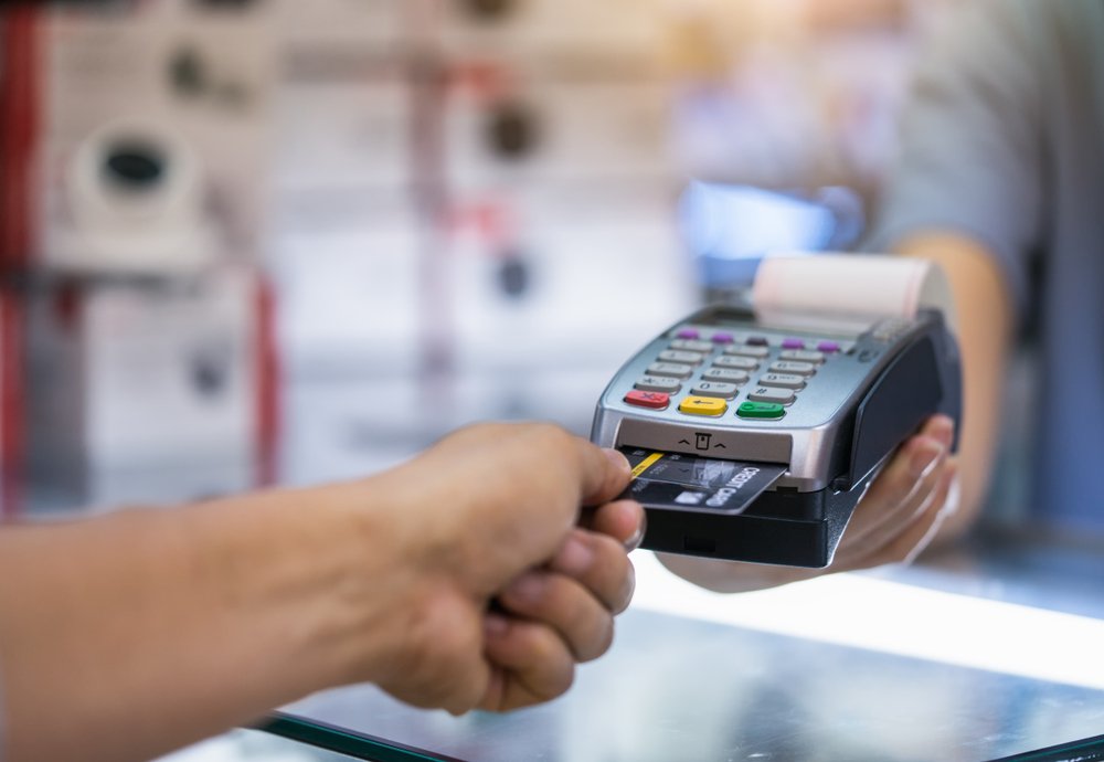 A mão de um homem branco inserindo um cartão de crédito em uma maquininha dentro de uma loja de eletrônicos
